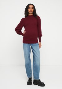 Kvinne står med hånden på hoften, iført en burgunder rippet genser, lyseblå jeans og svarte plattform støvler mot en hvit bakgrunn.