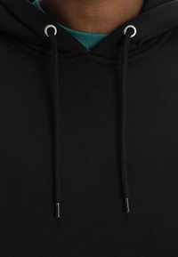 Sudadera negra con capucha y ojales plateados para los cordones. Presenta una textura suave y está superpuesta sobre una camiseta teal.