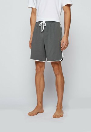 Mann trägt graue Shorts mit Kordelzug und weißem Besatz sowie ein weißes T-Shirt, steht barfuß vor einem schlichten hellen Hintergrund.