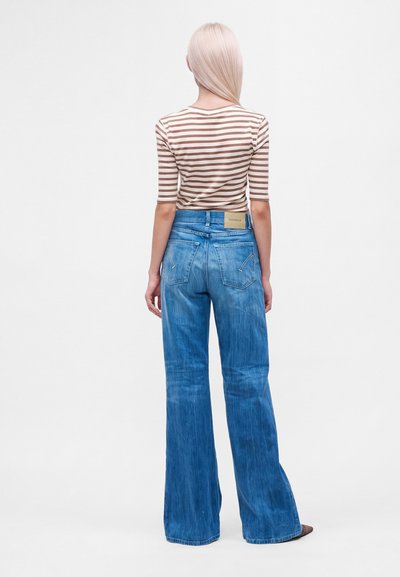 Femme aux cheveux blonds et raides portant un haut rayé marron et blanc et un jean bleu taille haute à jambes larges, debout de dos sur fond blanc.