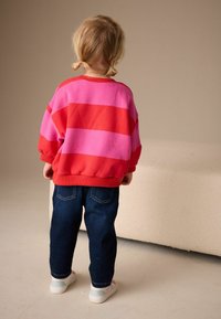 Sudadera de rayas rojas y rosas, de corte ancho, combinada con jeans azul oscuro y zapatillas blancas. Niño visto desde atrás.