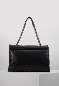 Černá kožená kabelka přes rameno s prošívaným horním okrajem a stříbrným řetízkovým popruhem, vystavená na bílém podstavci na světle šedém pozadí.