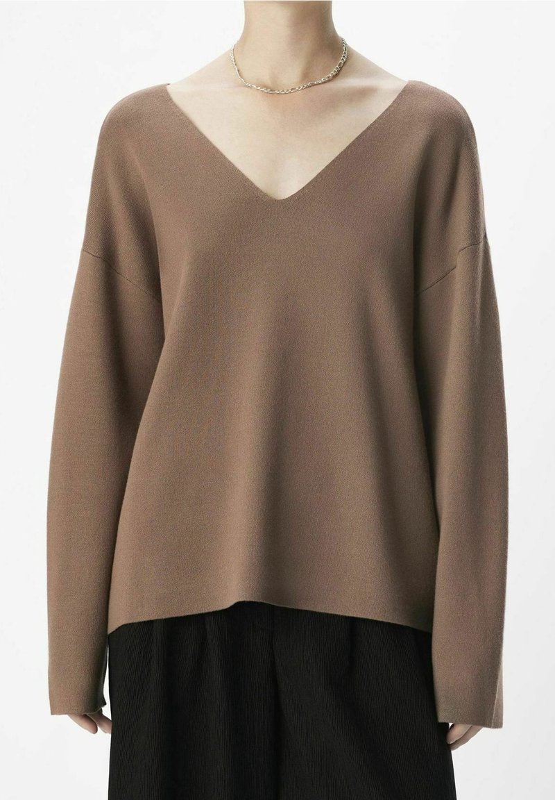 Personne portant un pull marron ample à col en V et un pantalon noir avec un collier chaîne argenté sur fond blanc.