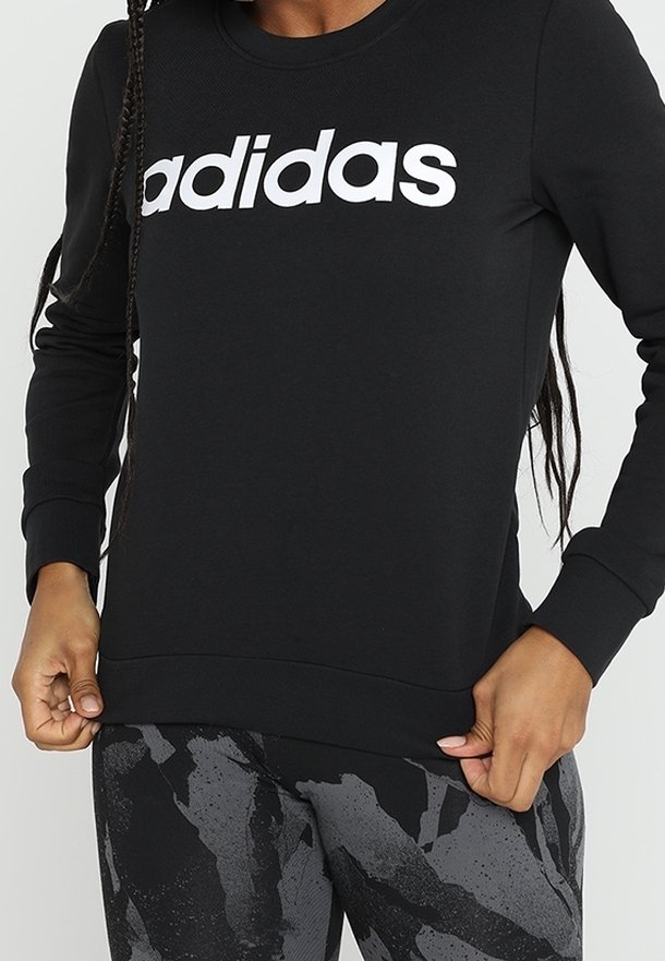 Sweatshirt noir avec le logo "adidas" blanc, manches longues, col rond et tissu texturé. Leggings noirs et gris à motifs complètent la tenue.