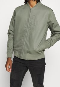 Bomber w kolorze oliwkowym, wykonany z gładkiego materiału, z zamkiem błyskawicznym z przodu, ściągaczami przy mankietach i detalem kieszeni z przodu. W połączeniu z czarnymi podartych dżinsami.