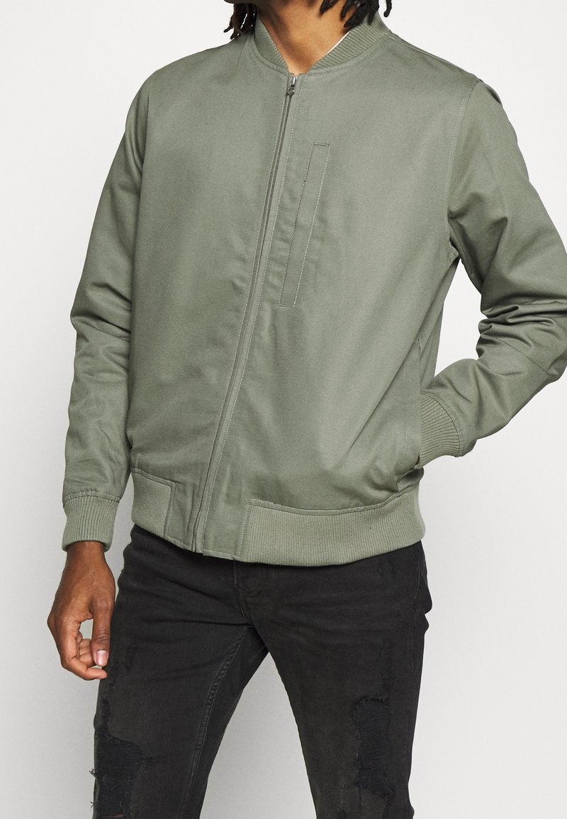 Bomber w kolorze oliwkowym, wykonany z gładkiego materiału, z zamkiem błyskawicznym z przodu, ściągaczami przy mankietach i detalem kieszeni z przodu. W połączeniu z czarnymi podartych dżinsami.