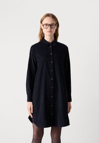 Donna che indossa un vestito camicia oversize nero con bottoni e maniche lunghe, con collant neri trasparenti, in piedi davanti a uno sfondo bianco semplice.