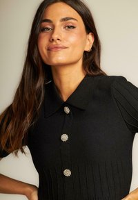 Top de punto negro con cuello clásico, que cuenta con tres botones metálicos decorativos y detalles de canalé vertical en el cuerpo y las mangas.