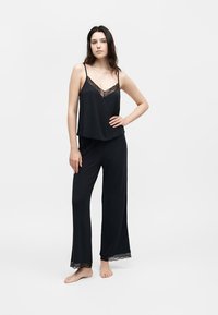 Νεαρή γυναίκα στέκεται ξυπόλυτη, φοράει μαύρο αμάνικο τοπ και φαρδιά παντελόνια με δαντέλα στο ντεκολτέ και στις άκρες των παντελονιών.