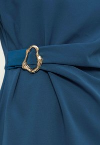 Ceinture en tissu bleu serrée par une boucle en métal doré irrégulière et brillante, créant des plis rassemblés dans le textile.