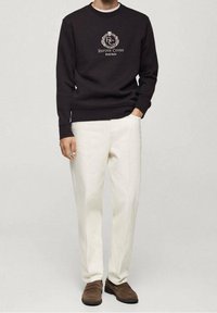 Sudadera negra con el logo "Refuge Cover", combinada con pantalones blancos. El conjunto presenta un ajuste relajado y zapatos casuales marrones.