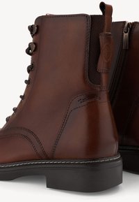 Stivali alla caviglia in pelle marrone con occhielli metallici, pannelli laterali testurizzati e una suola in gomma nera a contrasto. Presentano una cerniera laterale per un facile accesso.