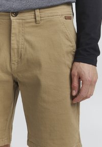 Shorts en coton beige avec une coupe classique, présentant une fermeture à bouton sur le devant, des poches latérales et une étiquette de marque discrète sur la taille.