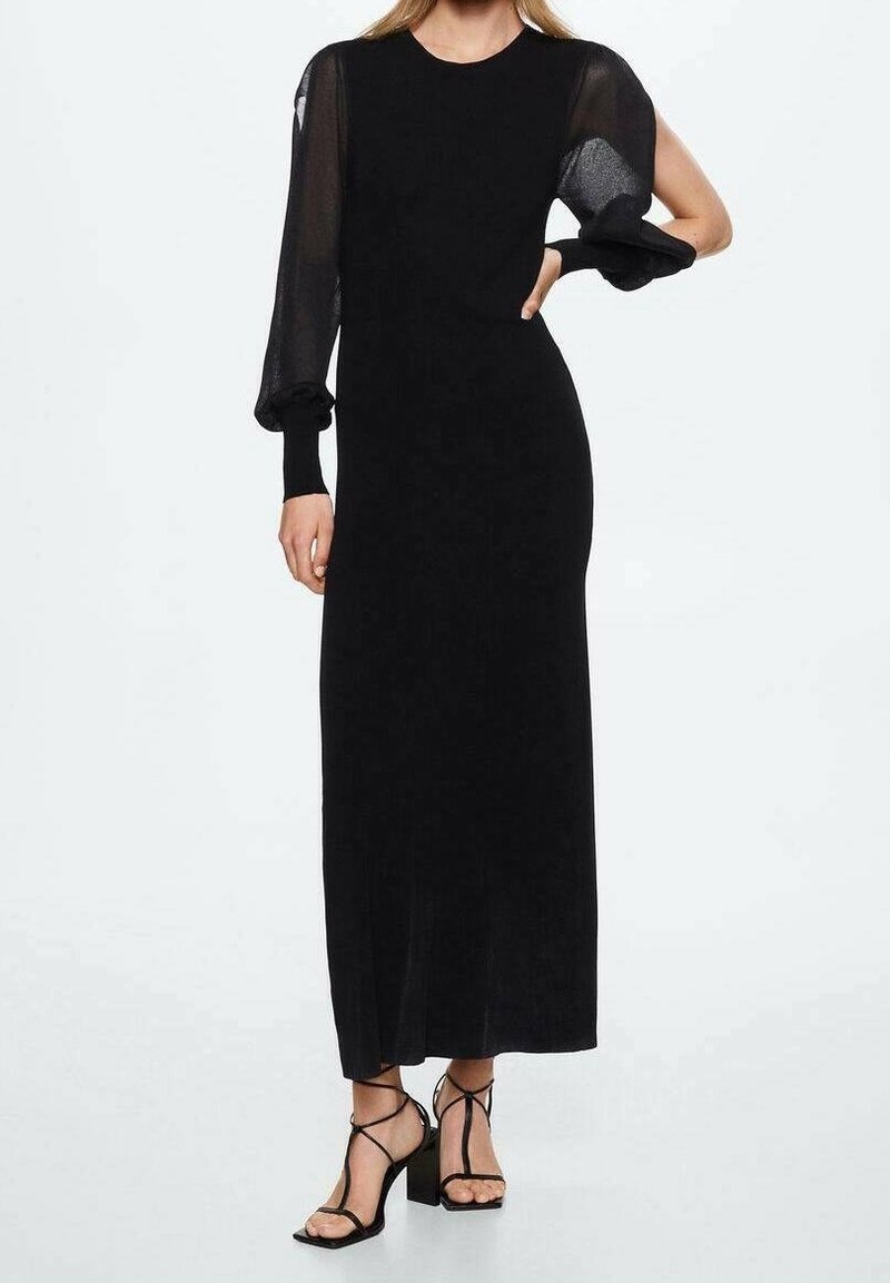 Femme portant une longue robe noire avec une manche bouffante transparente et des sandales à talons noirs avec brides à la cheville.