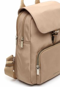 Beežist nailonist seljakott kaanega, kahe tõmblukuga taskuga ja kuldse tooniga käepidemetega. Omadusteks reguleeritavad rihmad ja sile tekstuur.