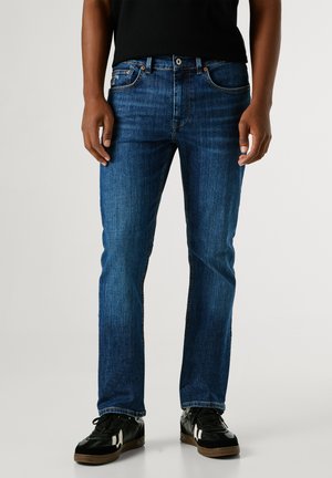 Dunkelblaue Jeans, gerade geschnitten, mit kontrastierendem orangefarbenem Nähen, Fünf-Taschen-Design und Metallknopfverschluss. Schwarze Sneaker runden den Look ab.