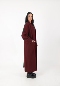 Abrigo largo burdeos con un corte relajado, mangas anchas y bolsillos laterales, hecho de una tela suave. Lleva puesto sobre una blusa negra, combinado con botas de plataforma.