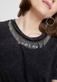Camiseta negra con un escote texturizado que presenta flecos metálicos y la palabra "REPLAY" en letras multicolores. Tejido suave y casual.
