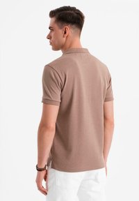 Hnědá polo košile vyrobená z bavlny, s krátkými rukávy, límečkem a bočními rozparky; nošena s bílými kalhotami, což vytváří čistý a minimalistický design.