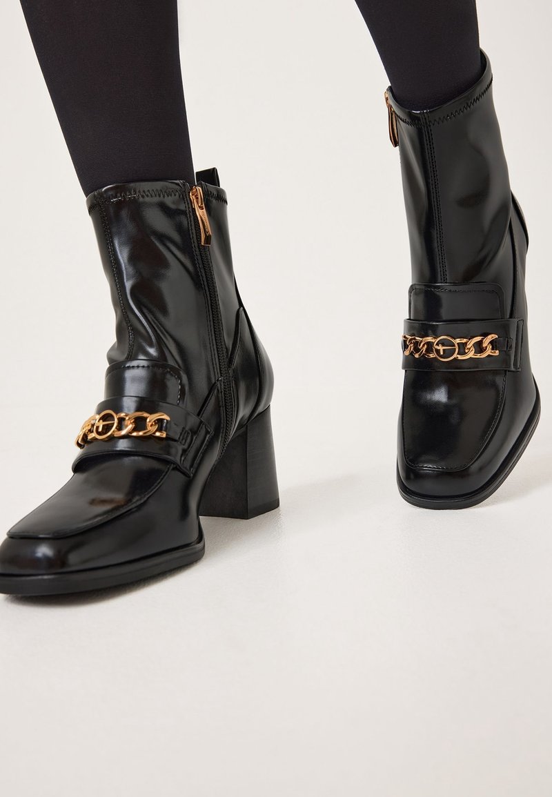 Bottines en cuir verni noir avec détail chaîne dorée, talons blocs et fermetures éclair latérales portées avec des collants noirs sur fond blanc.