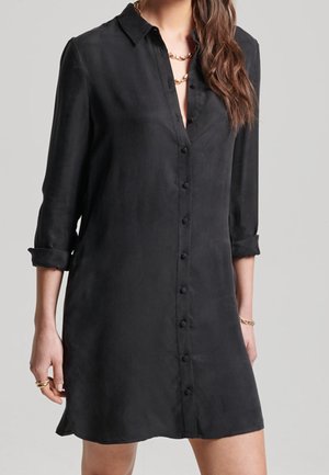 Vrouw draagt een zwarte overhemdjurk met opgerolde mouwen, geaccentueerd met een gouden ketting, armband en ringen, tegen een lichte achtergrond.