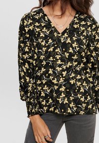 Svart blus med ett blommönster i gult och vitt. Har en V-ringning, långa ärmar med rynkade manschetter, och är i ett lätt tyg.