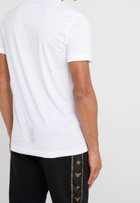 Bílé tričko s krátkým rukávem z hladké látky, se pruhovaným designovým prvkem na zádech, doplněné černými kalhotami se zlatými detaily.