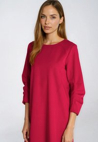 Młoda kobieta o długich, jasnobrązowych włosach, ubrana w prostą, długorękawową, ciemnoróżową sukienkę, stojąca na tle jednolitego jasnoszarego tła.