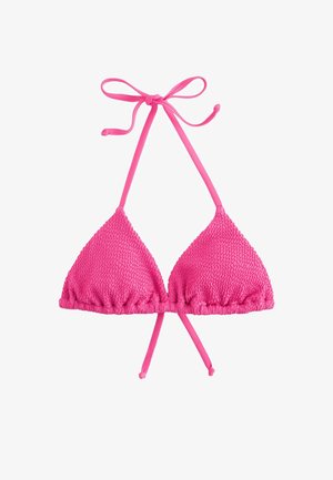 Jeune femme en bikini rose texturé debout sur une plage rocheuse avec des vagues de l'océan en arrière-plan sous un ciel dégagé.