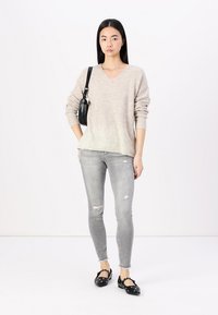 Beiger Strickpullover mit V-Ausschnitt und geripptem Muster, kombiniert mit hellgrauen, kaputten Jeans. Schwarze Ballerinas mit dekorativen Hardware-Elementen und einer kleinen schwarzen Tasche.