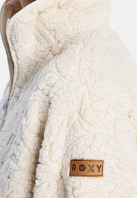 Damska kurtka polarowa w kolorze kremowym, z teksturowanego materiału z pikowanym wzorem, z zamkiem błyskawicznym oraz brązową naszywką marki Roxy.