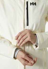 Witte soft-shell jas met een voorzak met rits, verstelbare mouwboorden en zichtbaar logo. Gladde textuur met klittenbandaccenten.