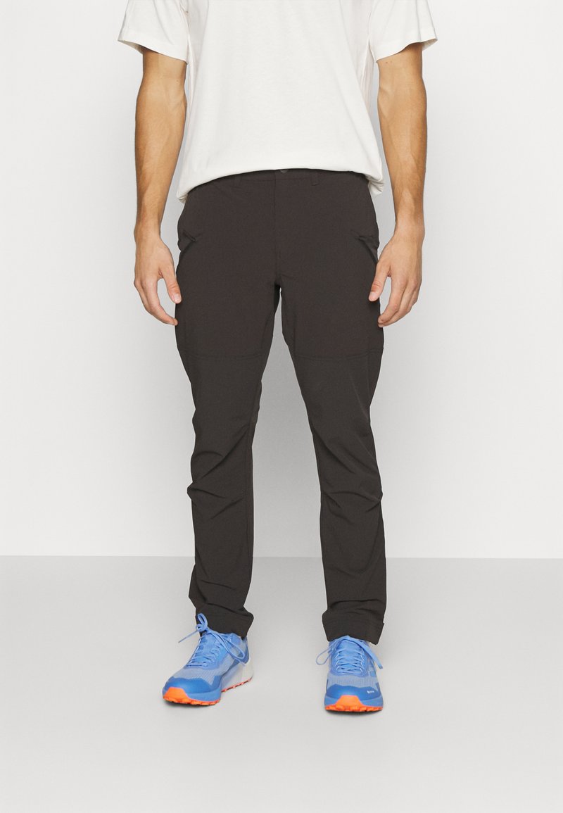 Črne lahke hlače s slim fit krojem, z zadrgo in gladko teksturo. V kombinaciji z modrimi športnimi čevlji z oranžnimi detajli.