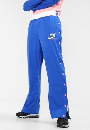 Personne portant un pantalon de sport bleu Nike avec des boutons latéraux roses et des baskets noires, debout les bras croisés sur un fond blanc.
