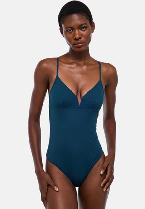 Femme aux cheveux courts portant un maillot de bain une pièce bleu sarcelle foncé avec des bretelles fines et un col en V, debout devant un fond uni.