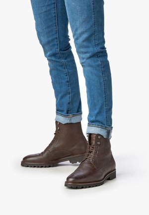 Botas de cuero marrón hasta el tobillo con una superficie texturada, punta redonda y suela resistente. Combinadas con jeans de denim azul remangados.