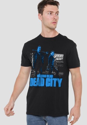 Man wearing black t-shirt with blue graphic of two characters holding weapons and text "Walking Dead Dead City" in a cityscape.