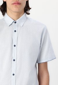 Camisa de botão de manga curta em tecido azul claro com uma textura subtil, apresentando um colarinho pontiagudo e botões escuros.