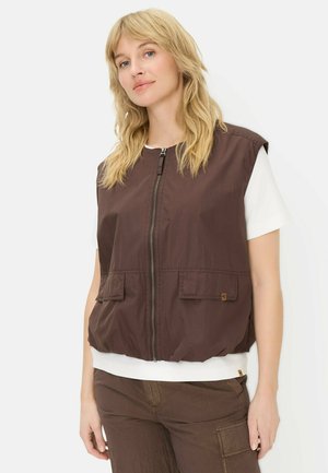 Woman with blonde hair wearing a brown zip-up vest over a white short-sleeve shirt and matching brown pants, standing against a white background.