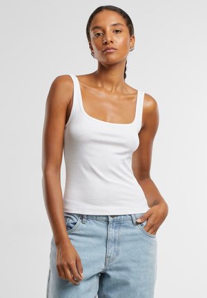 Kvinne med mørkt hår i hestehale iført hvit tanktop og lyseblå jeans med en hånd i lomma foran en ensfarget bakgrunn.