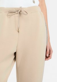 Pantalons de survêtement beiges avec une texture douce. Comprend une taille élastique avec cordon de serrage et des accents métalliques sur le côté.