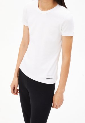 Kobieta ubrana w biały prążkowany T-shirt z krótkim rękawem z nadrukiem "ARMEDANGELS" oraz czarne dopasowane spodnie, stojąca na białym tle.