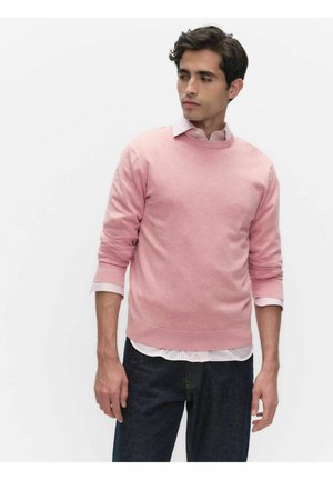 Man met een lichtroze trui over een wit overhemd met kraag en donkere spijkerbroek, staand tegen een effen witte achtergrond, kijkend naar de zijkant.