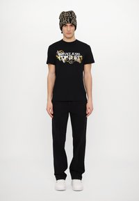 Juodas medvilnės marškinėliai su balta ir auksine "Versace Jeans Couture" grafika, derinami su juodomis kelnėmis ir juodu bei auksiniu raštuotu kepurės.
