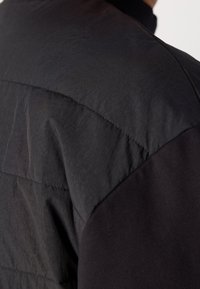 Gros plan sur l'épaule d'une veste matelassée noire avec des lignes horizontales cousues et une texture de tissu lisse et mate.