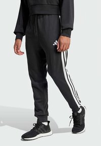 Μαύρο παντελόνι φόρμας με λευκές ρίγες στο πλάι, ελαστική μέση και τελείωμα με μανσέτες, σε συνδυασμό με μαύρα αθλητικά παπούτσια με σχεδιασμό από δίχτυ.