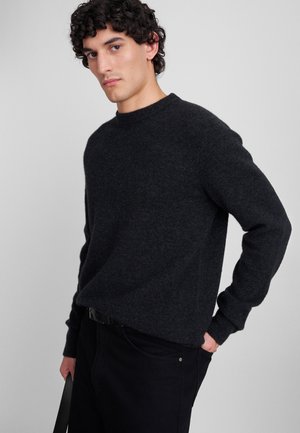 Donkergrijze wollen trui met een ronde hals, geribbelde mouwen en zoom. Het model draagt zwarte jeans en houdt in één hand een riem vast.