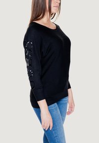 Maglione nero con una vestibilità ampia, maniche a 3/4 e dettagli texture sulla manica sinistra. Abbinato a jeans in denim blu chiaro.