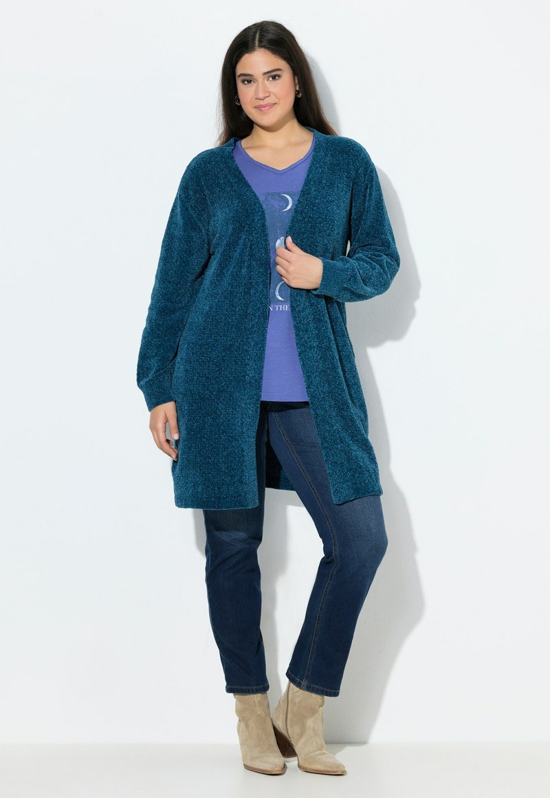 Turkos fluffig cardigan med långa ärmar, öppen framsida och sidslitsar, lager-på-lager över en lila grafisk t-shirt och matchad med mörka jeans och beige ankelstövlar.