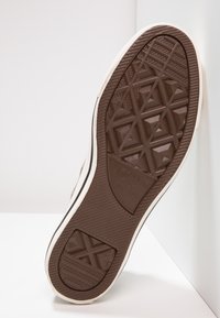 Semelle de sneaker en caoutchouc brun avec un motif texturé, mettant en avant des formes géométriques et un logo central. Les bords sont de couleur crème avec une finition lisse.